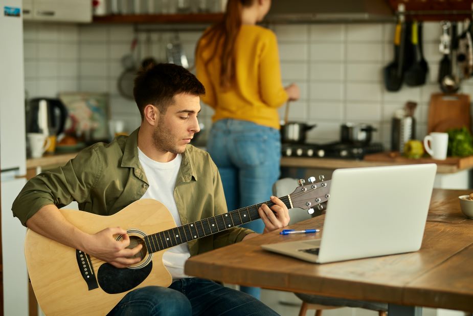 Joven disfrutando de su hobby tocando la guitarra frente a su computadora.